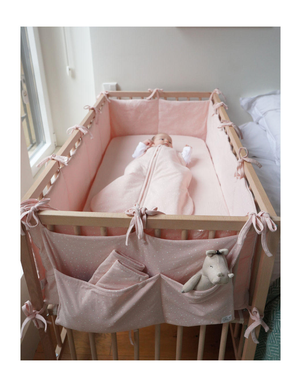 Organizador de cuna rosa con bolsillos para bebé instalado en la cuna, práctico accesorio para guardar pañales, chupetes y productos de cuidado infantil.